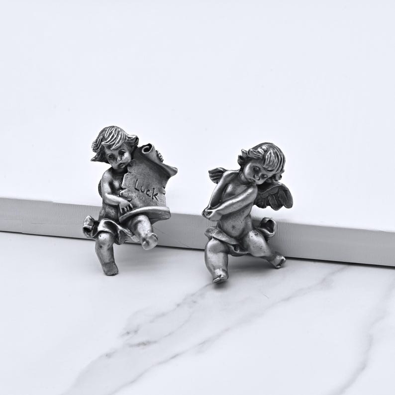 Peut inclure: Deux figurines de ch&eacute;rubins argent&eacute;s. L'un tient un parchemin avec le mot "Luck". L'autre ch&eacute;rubin a des ailes et est agenouill&eacute;. Les figurines sont d&eacute;taill&eacute;es et ont une esth&eacute;tique vintage.