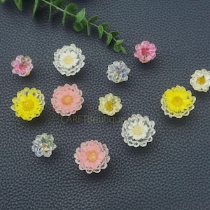 Może przedstawiać: Kolekcja kolorowych kaboszonów kwiatowych w żywicy. Kwiaty są w różnych kolorach, w tym żółtym, różowym, białym i niebieskim. Kaboszony ułożone są na ciemnoszarej powierzchni, z gałązką zielonych liści w tle.