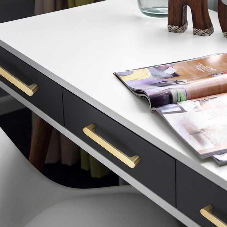 May include: A contemporary desk featuring a white surface and three black drawers. The drawers are fitted with gold-coloured, rectangular handles. An open magazine lies on the desk. Suitable for a home office or workspace.