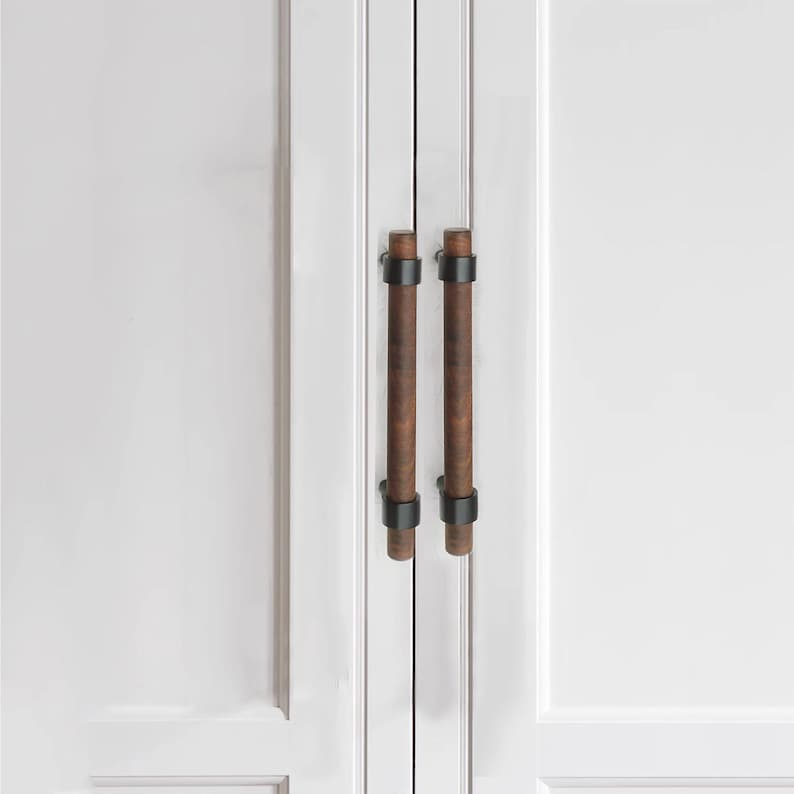 以下が含まれることがあります： 白いキャビネットドアのクローズアップ。2つの垂直な円筒形のキャビネットプルがあります。プルは濃い茶色の木材と黒い金属のアクセントで作られています。キャビネットドアは2つのパネルに分かれています。