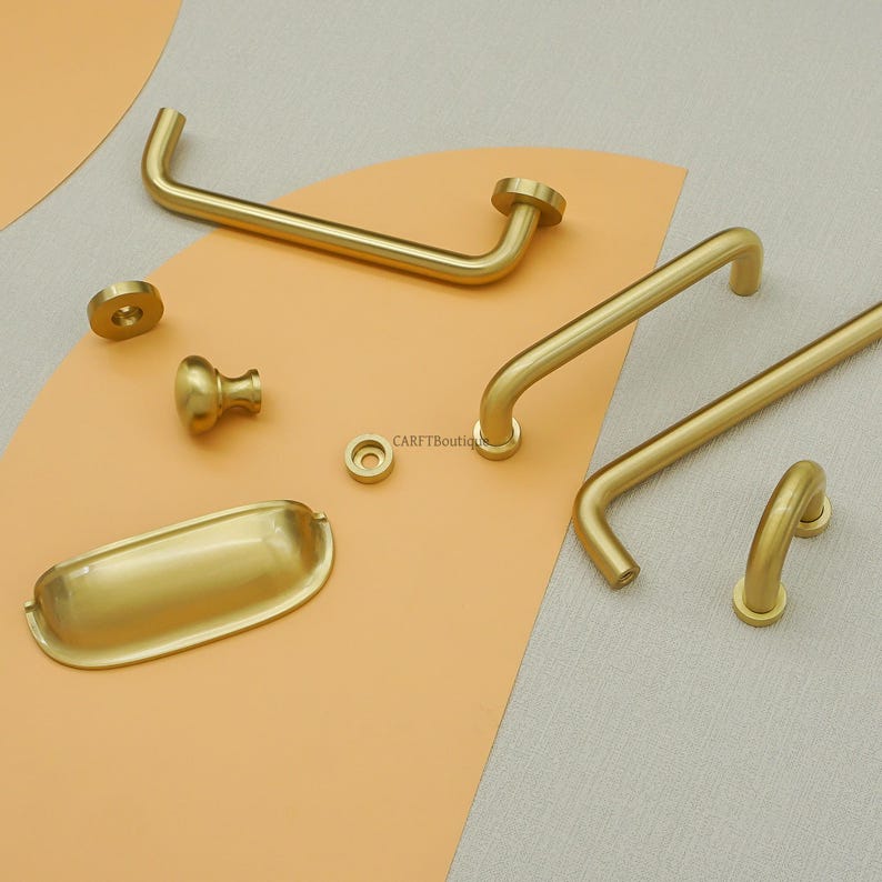 May include: A selection of gold-coloured cabinet hardware, including handles, knobs, and a pull. The hardware pieces are arranged on a neutral background, showcasing their various shapes and designs. The hardware is made of metal.