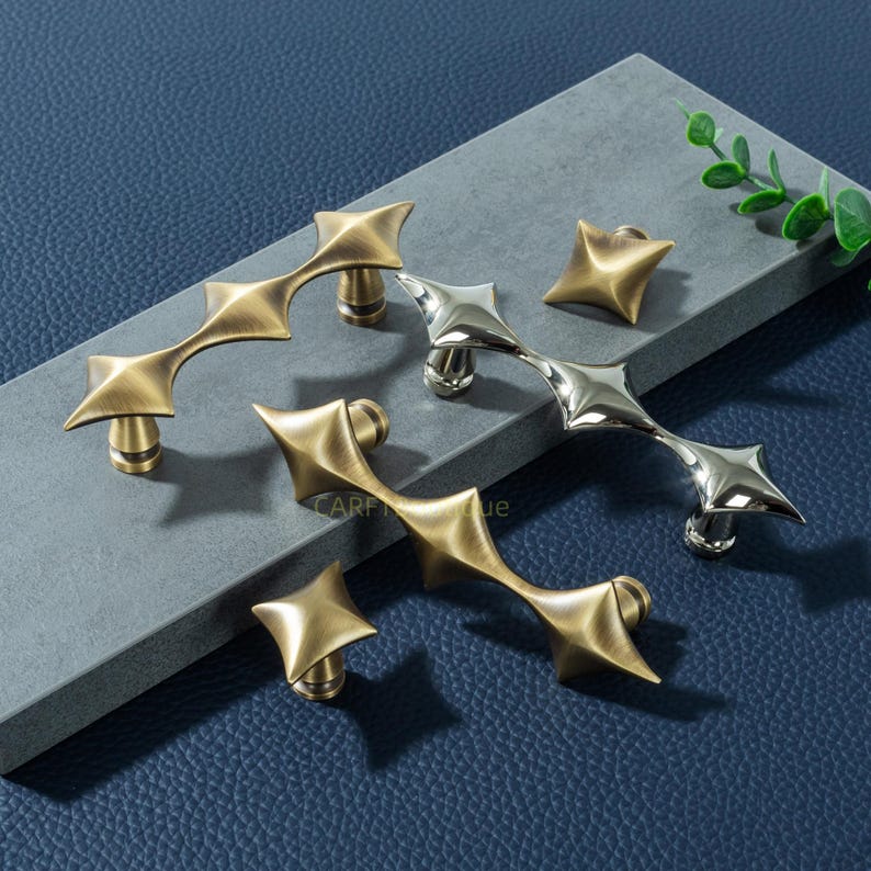 以下が含まれることがあります： 星型のノブとプルを含む、装飾的なキャビネットハードウェアのコレクション。 プルは真鍮仕上げのユニークな幾何学模様のデザインで、1つのプルとノブはシルバー仕上げです。 青い背景に対して灰色の表面に表示されます。