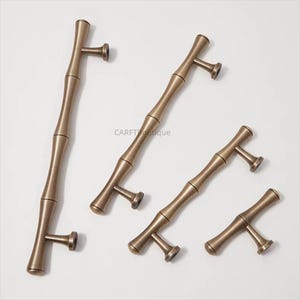 K&ouml;nnte beinhalten: Bronzefarbene Schrankgriffe mit Bambus-Design. Die Griffe haben eine zylindrische Mitte mit ausgestellten Enden und einen T-f&ouml;rmigen Griff. Die Hardware ist auf einer wei&szlig;en Oberfl&auml;che angeordnet. Der Text "CARFTboutique" ist sichtbar.