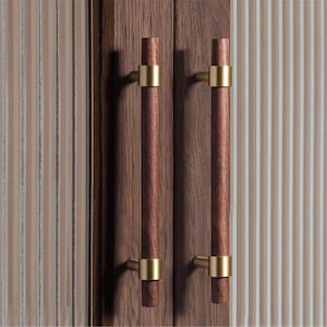 以下が含まれることがあります： 2つの縦型木製ハンドルが付いたキャビネットドアのクローズアップ。ハンドルは濃い茶色で、金色の金属アクセントが付いています。キャビネットドアは濃い茶色の木材で、背景は薄茶色と白のストライプ模様です。