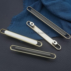 Peut inclure: Plusieurs poignées de meubles de différents modèles. Les poignées ont un centre rectangulaire noir, blanc ou gris, encadré par une poignée en métal doré ou argenté. Elles sont présentées sur une surface bleu foncé.