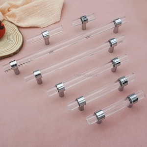 May include: Clear acrylic cabinet pulls with polished chrome hardware. The pulls are cylindrical and have a modern design. The hardware is silver and connects the acrylic rods to the cabinet. The pulls are arranged on a pink surface.