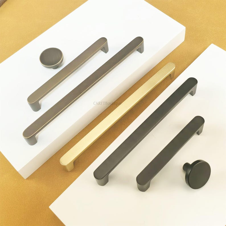 May include: A selection of cabinet handles and knobs in bronze, gold, and black finishes. The image shows long bar pulls and matching round knobs. The hardware is displayed on a white surface.
