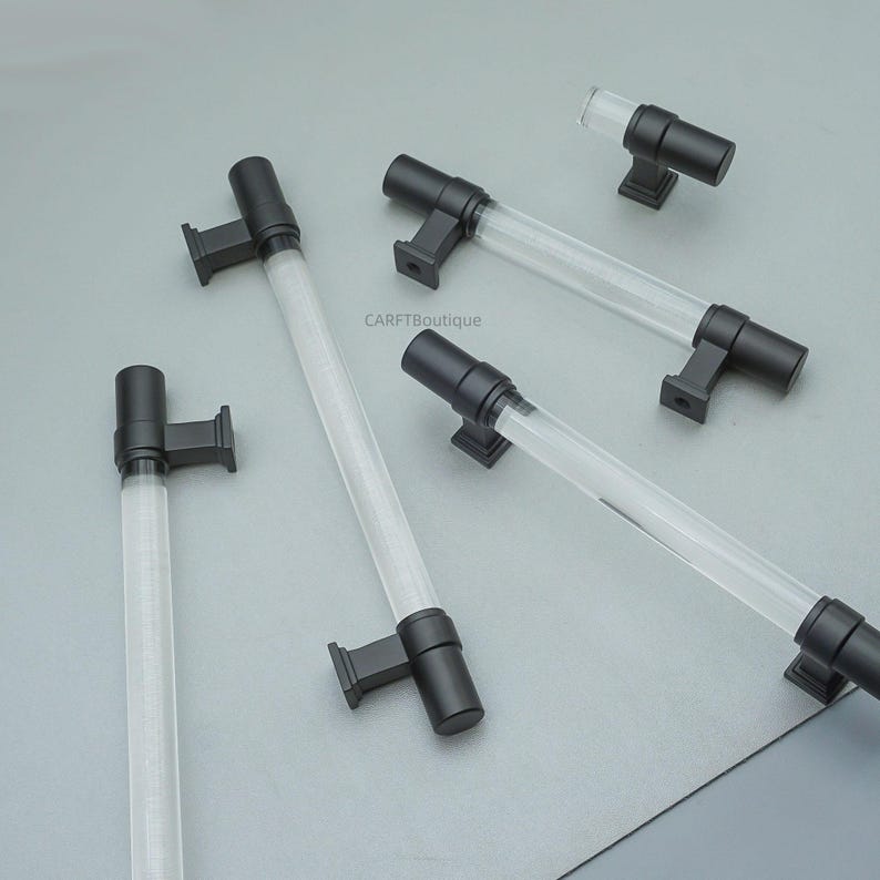 K&ouml;nnte beinhalten: Mehrere Schrankgriffe mit einem transparenten, zylindrischen Mittelteil und schwarzer Hardware. Die Griffe sind auf einer grauen Oberfl&auml;che angeordnet. Der transparente Mittelteil scheint aus Acryl oder Glas zu bestehen. Die Hardware ist quadratisch und rechteckig.