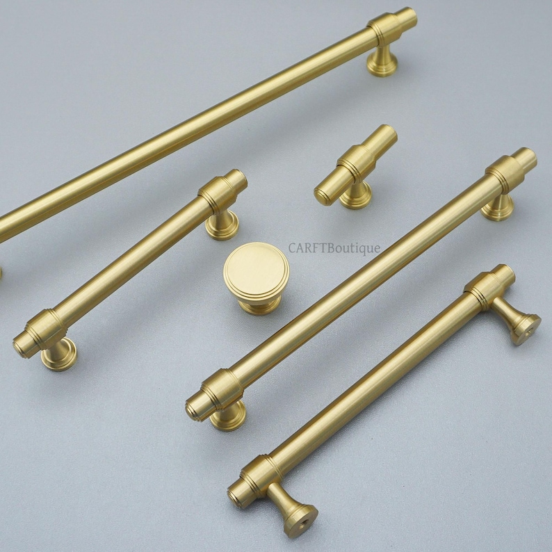 K&ouml;nnte beinhalten: Eine Sammlung goldfarbener Schrankbeschl&auml;ge, darunter mehrere Stangengriffe und ein runder Knopf. Die Beschl&auml;ge haben ein zylindrisches Design mit dekorativen Details und einer matten Oberfl&auml;che. Die Teile sind auf einer hellgrauen Oberfl&auml;che angeordnet.