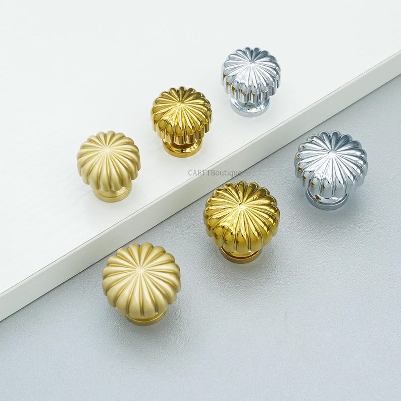 May include: A collection of decorative cabinet knobs in gold and silver finishes. The knobs have a fluted, flower-like design. The image is taken from a high angle, showcasing the hardware against a neutral background. The brand name "CARE Boutique" is visible.