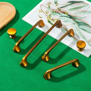 Pode incluir: Uma coleção de ferragens de armário de latão antigo, incluindo dois botões e três tamanhos diferentes de puxadores. As ferragens estão dispostas sobre uma superfície verde, com uma bandeja de madeira e uma revista ao fundo.