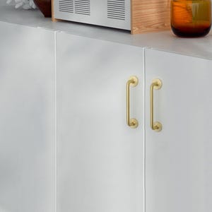May include: Close-up of a white cabinet with two gold-colored, curved handles. The handles are vertically oriented and have a simple, elegant design. The cabinet is part of a kitchen or storage unit.