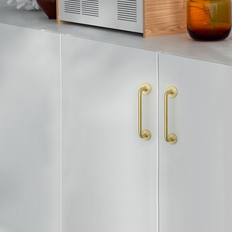 May include: Close-up of a white cabinet with two gold-colored, curved handles. The handles are vertically oriented and have a simple, elegant design. The cabinet is part of a kitchen or storage unit.