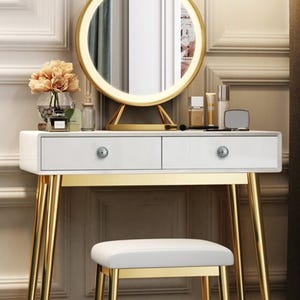May include: A white vanity table with gold legs and a matching stool. The table has two drawers with decorative knobs and a round, illuminated mirror. A vase of flowers and beauty products are displayed on the table.