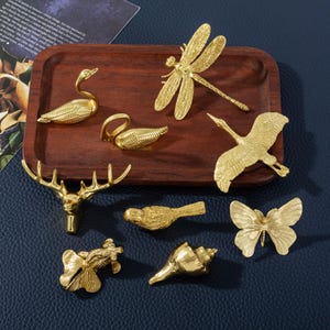 May include: A selection of gold-coloured decorative figurines, including a swan, dragonfly, bird, butterfly, deer head, fish, and seashell, displayed on a wooden tray. The figurines are detailed and have a metallic finish. The tray is on a blue surface.