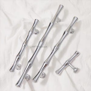 K&ouml;nnte beinhalten: Eine Sammlung gl&auml;nzender, silberfarbener Schrankgriffe und -kn&ouml;pfe im Bambus-Stil. Die Hardware ist auf einer wei&szlig;en Oberfl&auml;che angeordnet, mit dem Markennamen "CARFTboutique" sichtbar. Die Griffe haben ein schlankes, modernes Design.