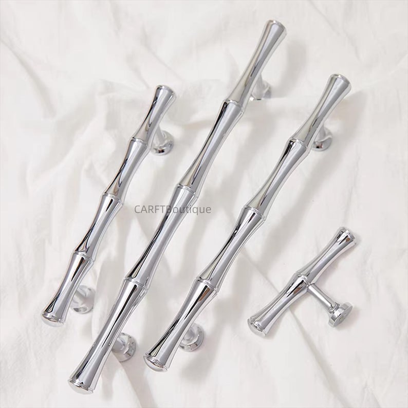 K&ouml;nnte beinhalten: Eine Sammlung gl&auml;nzender, silberfarbener Schrankgriffe und -kn&ouml;pfe im Bambus-Stil. Die Hardware ist auf einer wei&szlig;en Oberfl&auml;che angeordnet, mit dem Markennamen "CARFTboutique" sichtbar. Die Griffe haben ein schlankes, modernes Design.