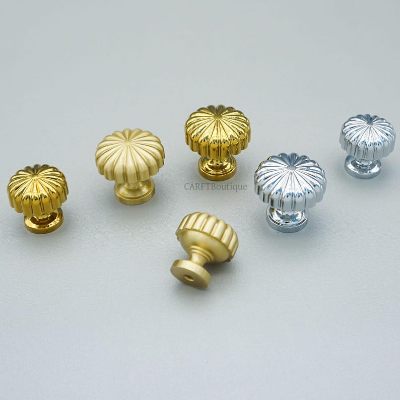 May include: A collection of decorative cabinet knobs in various metallic finishes. The knobs feature a fluted, flower-like design. Colors include gold, silver, and a muted brass tone. The knobs are shown on a light gray surface.