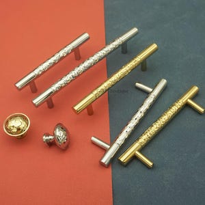 May include: An assortment of cabinet hardware, including silver and gold-toned handles and knobs. The handles have a textured, hammered design, while the knobs feature a round, hammered surface. The hardware is displayed on a red and blue-grey background.