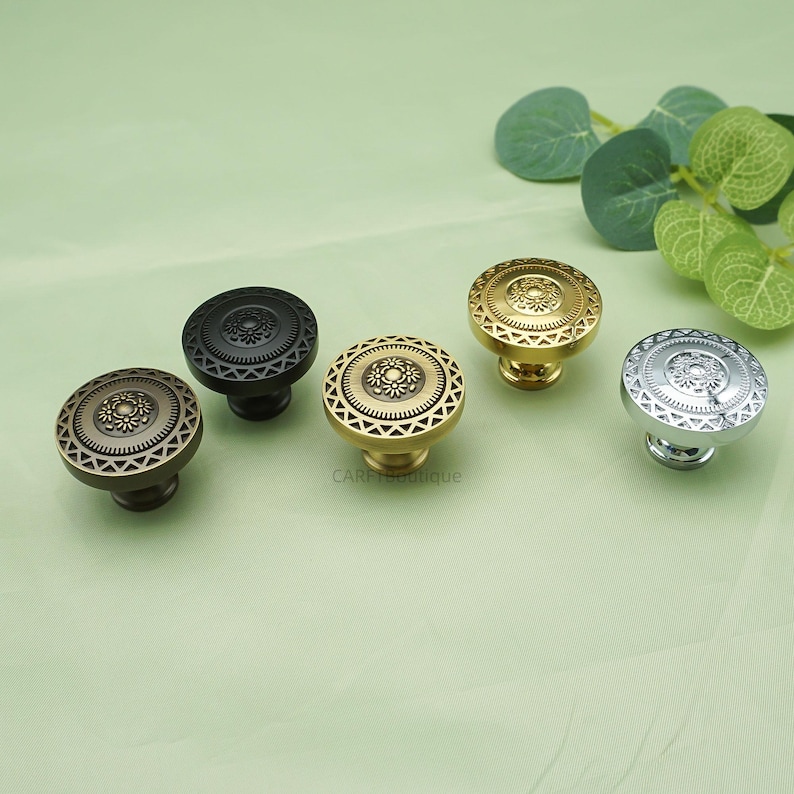 May include: Five decorative cabinet knobs in various metallic finishes: bronze, black, gold, and silver. Each knob features a circular design with a central floral pattern and a geometric border. The knobs are arranged on a light green surface.