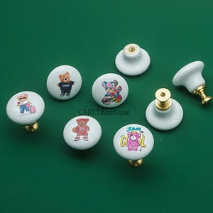 May include: A collection of white ceramic cabinet knobs with gold-toned hardware. Each knob features a different cartoon bear design, including bears in outfits and floral patterns. The knobs are arranged on a green surface.