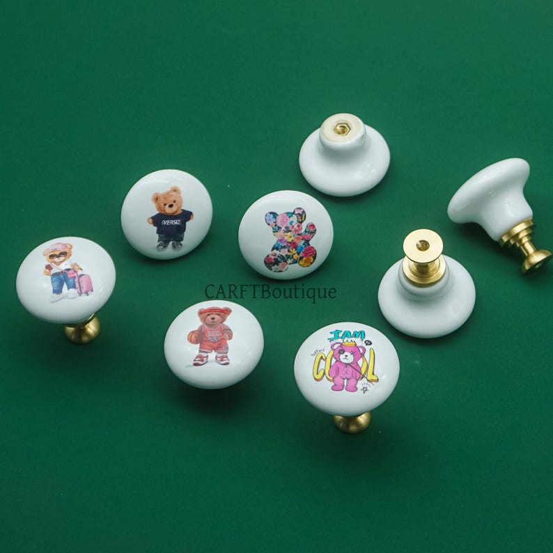 May include: A collection of white ceramic cabinet knobs with gold-toned hardware. Each knob features a different cartoon bear design, including bears in outfits and floral patterns. The knobs are arranged on a green surface.