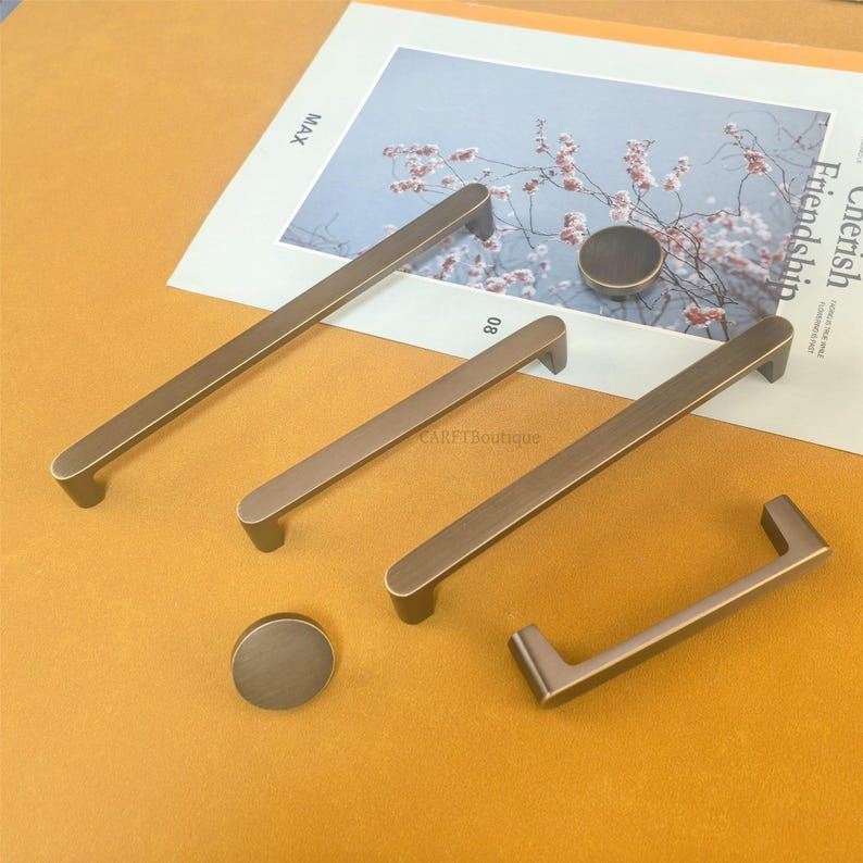 May include: A set of bronze-coloured cabinet handles and knobs. The set includes rectangular bar pulls and round knobs. The hardware is arranged on a light brown surface, with a magazine featuring floral imagery in the background. The hardware has a modern, minimalist design.