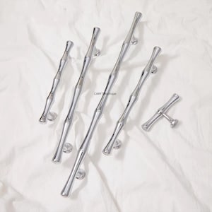 K&ouml;nnte beinhalten: Eine Sammlung von polierten silbernen Schrankgriffen und einem Knauf, alle im Bambus-Stil gestaltet. Die Beschl&auml;ge sind auf einem wei&szlig;en Stoffhintergrund angeordnet, der die reflektierende Qualit&auml;t des Metalls hervorhebt. Die Griffe sind unterschiedlich gro&szlig;.