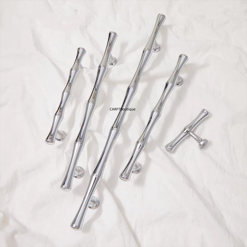 K&ouml;nnte beinhalten: Eine Sammlung von polierten silbernen Schrankgriffen und einem Knauf, alle im Bambus-Stil gestaltet. Die Beschl&auml;ge sind auf einem wei&szlig;en Stoffhintergrund angeordnet, der die reflektierende Qualit&auml;t des Metalls hervorhebt. Die Griffe sind unterschiedlich gro&szlig;.