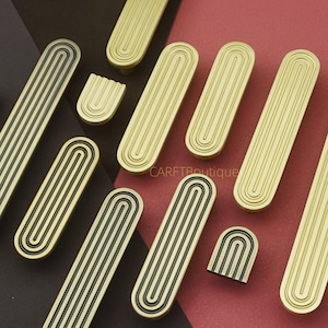 Puede incluir: Una colecci&oacute;n de tiradores y pomos dorados y negros para armarios. Los tiradores son largos, rectangulares con ranuras verticales y un borde negro. Los pomos son arqueados con ranuras verticales.