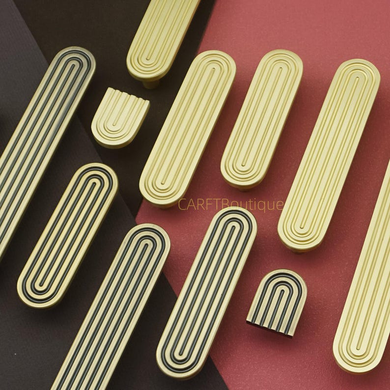 Puede incluir: Una colecci&oacute;n de tiradores y pomos dorados y negros para armarios. Los tiradores son largos, rectangulares con ranuras verticales y un borde negro. Los pomos son arqueados con ranuras verticales.