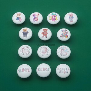 May include: A collection of white ceramic cabinet knobs with various designs. Some knobs feature teddy bears in different outfits, while others display unicorns and the words "LOVE", "PEACE", and "BE LIKE A UNICORN".