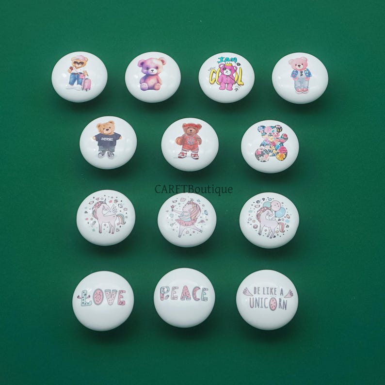 May include: A collection of white ceramic cabinet knobs with various designs. Some knobs feature teddy bears in different outfits, while others display unicorns and the words "LOVE", "PEACE", and "BE LIKE A UNICORN".