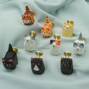 May include: Nine wooden cat-shaped cabinet knobs, hand-carved and painted in various colours. Designs include orange tiger stripes, black, white, and calico patterns. Each knob has a brass-coloured base. The text "CARFTBoutique" is visible.