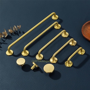 May include: A collection of brass-colored cabinet hardware, including bar pulls and round knobs. The bar pulls vary in length, with curved ends and circular mounting plates. The knobs are circular with a short stem. The hardware is displayed on a dark blue surface.
