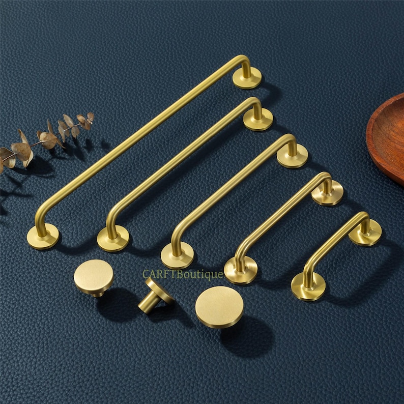 May include: A collection of brass-colored cabinet hardware, including bar pulls and round knobs. The bar pulls vary in length, with curved ends and circular mounting plates. The knobs are circular with a short stem. The hardware is displayed on a dark blue surface.