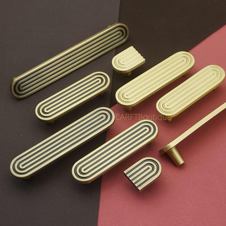 Puede incluir: Un surtido de tiradores y pomos de armario en tono lat&oacute;n. Los herrajes presentan un dise&ntilde;o ovalado acanalado con un acento oscuro. Se muestran varios tama&ntilde;os sobre un fondo marr&oacute;n y rosa.