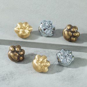 Puede incluir: Seis tiradores decorativos con forma de huella de pata en acabados dorado, plateado y bronce. Los tiradores están dispuestos sobre una superficie gris claro, mostrando su brillo metálico y las detalladas huellas de las almohadillas. Ideal para añadir fantasía.