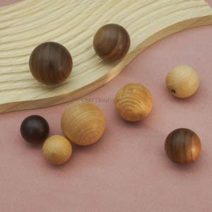May include: A collection of wooden spheres in various sizes and wood tones, including dark walnut and light pine. The spheres are arranged on a light pink surface and a wooden plank.