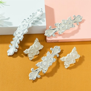 May include: White and silver decorative cabinet hardware. The hardware includes floral designs and ornate scrollwork. The image shows several pieces of hardware, including long pulls and smaller knobs. The hardware is displayed on a white, pink, and orange background.