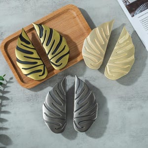 May include: A collection of decorative cabinet pulls in various metallic finishes. The pulls are leaf-shaped, with detailed veining. Finishes include gold with black accents, brushed gold, and a silver-gray. The pulls are displayed on a wooden tray and a gray surface.