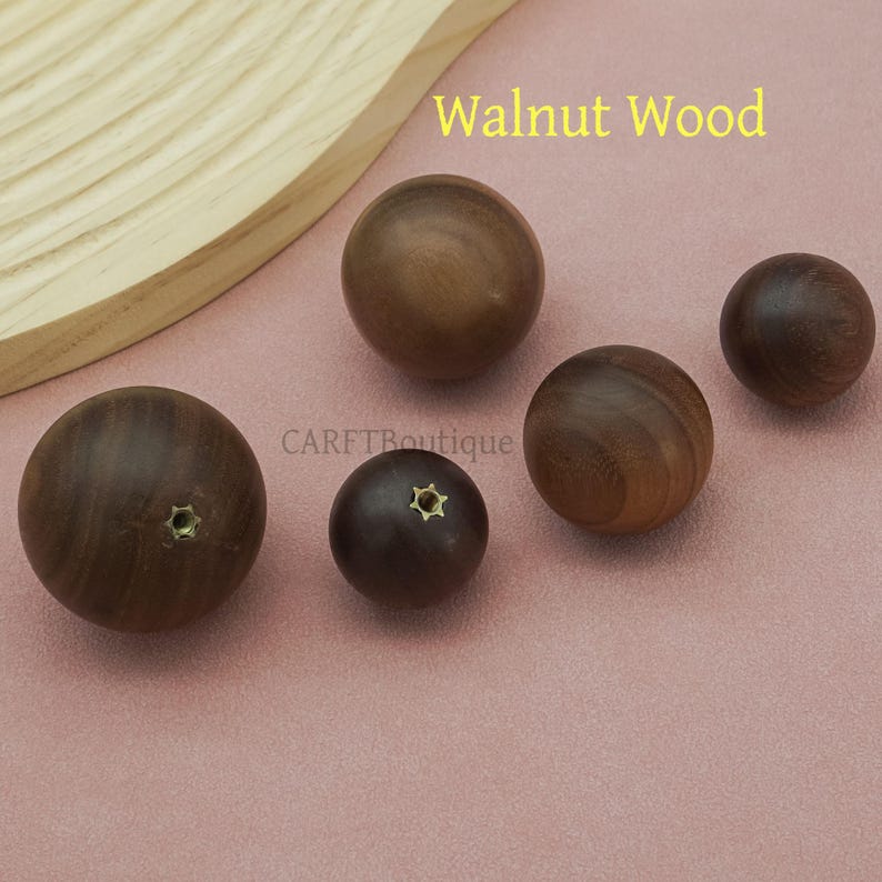 May include: A collection of walnut wood spheres in varying sizes. Some spheres have a small, star-shaped metal insert. The text "Walnut Wood" is displayed in yellow. The spheres are arranged on a pink surface.
