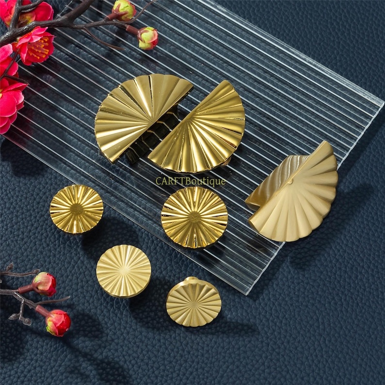 May include: A selection of gold-coloured cabinet hardware, including fan-shaped pulls and round knobs. The hardware is arranged on a clear, ridged surface, with a dark blue background. The image also includes red and pink flowers.
