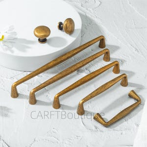 May include: A collection of antique brass cabinet hardware, including knobs and pull handles. The knobs are round and the handles are various lengths, all with a distressed finish. The hardware is arranged on a white surface.