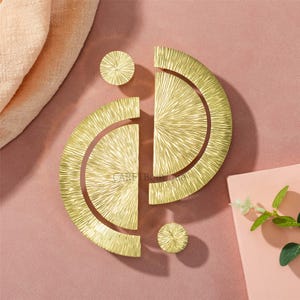 May include: Gold-colored cabinet hardware set, including two semi-circular handles and two round knobs. The hardware has a textured, radial design. The items are arranged on a pink surface with a peach-colored fabric and a pink book.