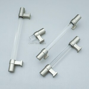 K&ouml;nnte beinhalten: Vier Schrankgriffe mit transparenten Acrylst&auml;ben und Hardware mit geb&uuml;rsteter Nickeloberfl&auml;che. Die Griffe haben ein modernes Design mit zylindrischer Stange und quadratischen Montageplatten. Die Hardware hat eine matte Oberfl&auml;che.