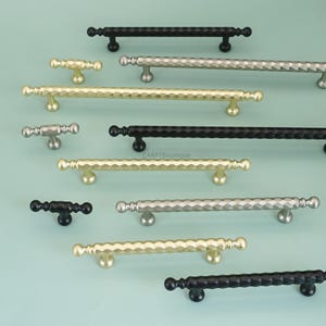 May include: A selection of cabinet handles and knobs in gold, silver, and black finishes. The hardware has a geometric, textured design. The items are displayed against a light blue background. Measurements are in centimetres.