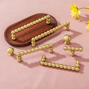 May include: A collection of gold-colored cabinet hardware, including handles and knobs, arranged on a pink surface. The handles feature a beaded design, and the knobs are spherical. A wooden tray and yellow flowers are also in the image.