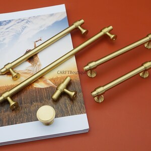 Pode incluir: Uma cole&ccedil;&atilde;o de ferragens douradas para arm&aacute;rios, incluindo puxadores de barra, um puxador em forma de T e um bot&atilde;o redondo. O hardware &eacute; exibido em uma superf&iacute;cie branca e impressa contra um fundo marrom avermelhado. O texto "CARFTBoutique" &eacute; vis&iacute;vel.