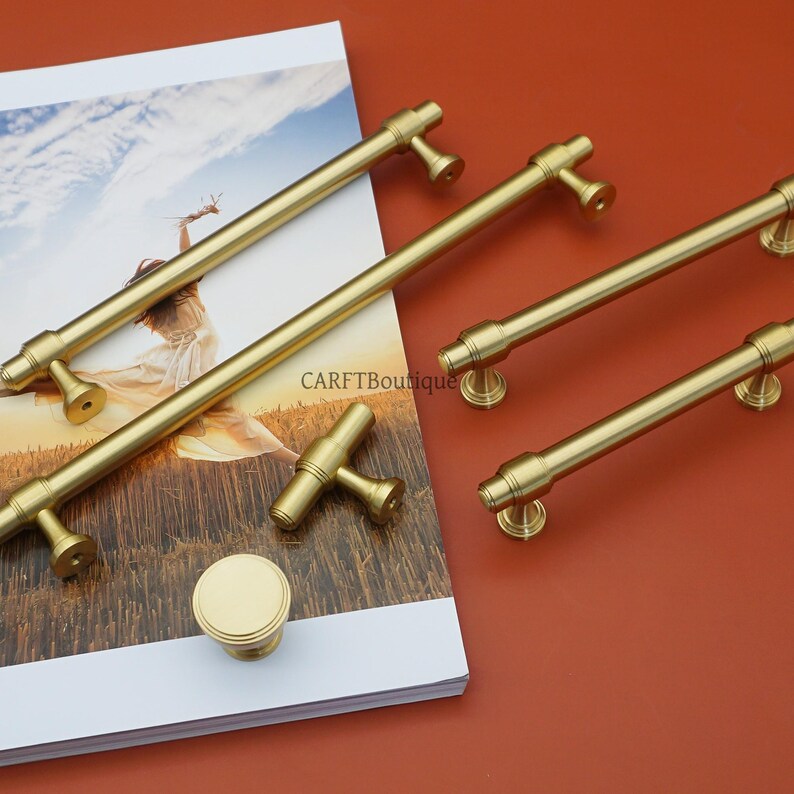 Pode incluir: Uma cole&ccedil;&atilde;o de ferragens douradas para arm&aacute;rios, incluindo puxadores de barra, um puxador em forma de T e um bot&atilde;o redondo. O hardware &eacute; exibido em uma superf&iacute;cie branca e impressa contra um fundo marrom avermelhado. O texto "CARFTBoutique" &eacute; vis&iacute;vel.